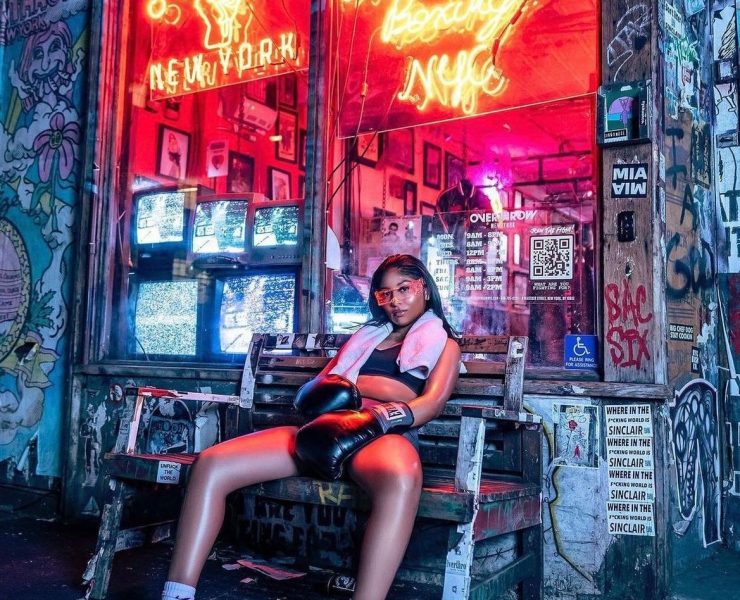 woman fighter with black hair sitting on a bench outside of a studio with a neon sigh that says Overthrow Boxing