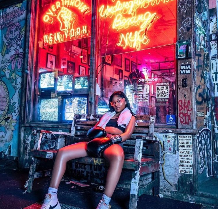 woman fighter with black hair sitting on a bench outside of a studio with a neon sigh that says Overthrow Boxing